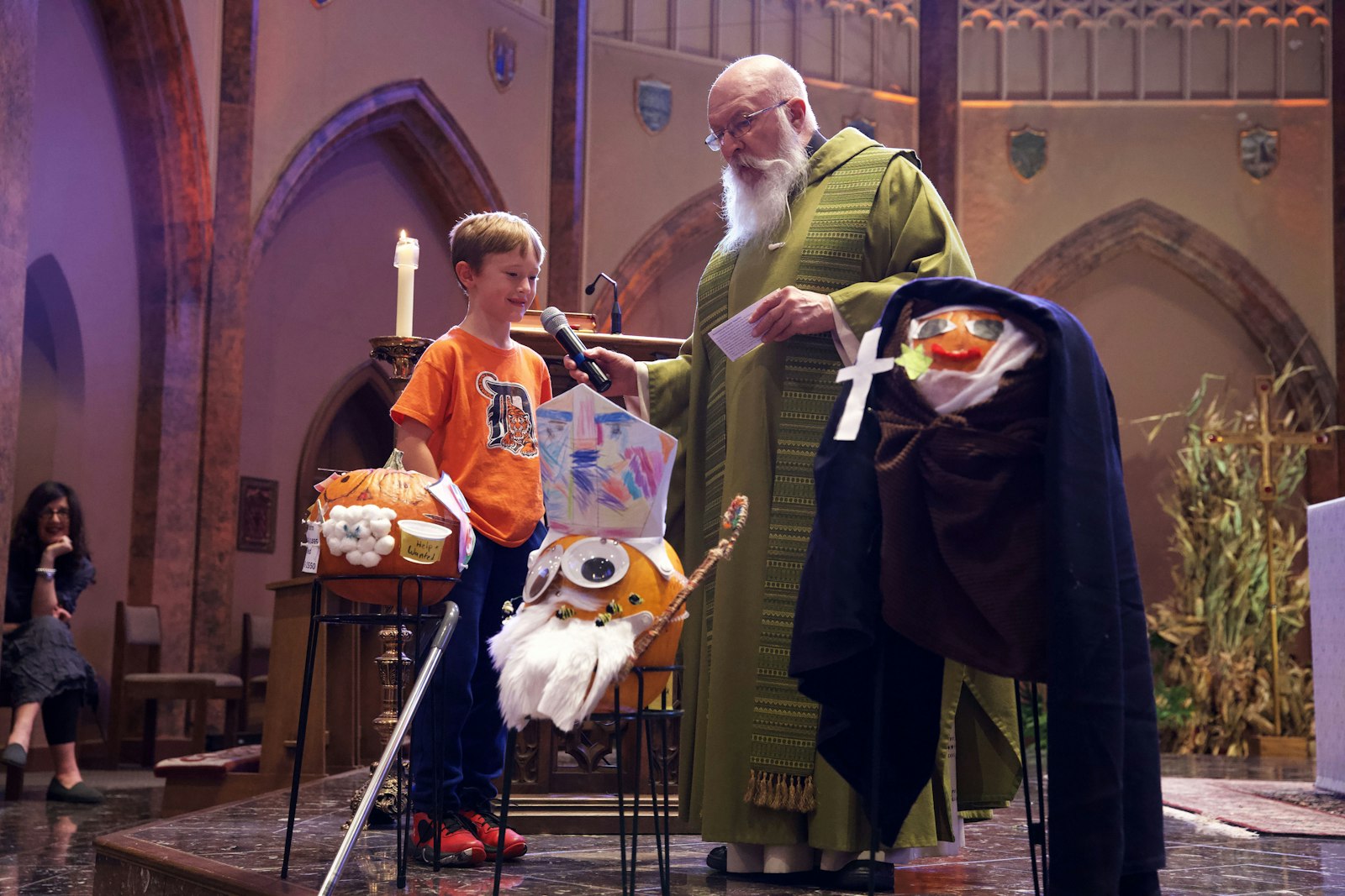 A kindergarten student tells Fr. Tim Pelc about St. Ambrose (center). The students also decorated pumpkins to look like St. John of God (left) and St. Edith Stein (right).