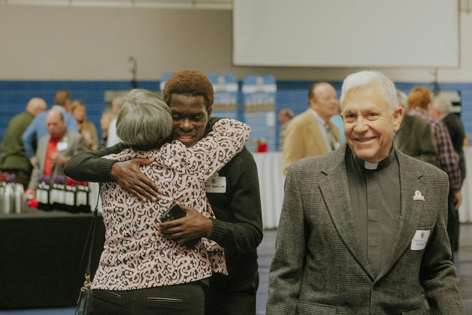 El hermano David Basil, OSB, recibe un abrazo de un simpatizante durante el evento de celebración de Catholic Central el 19 de noviembre mientras el p. Maurice Restivo, CSB, sonríe.