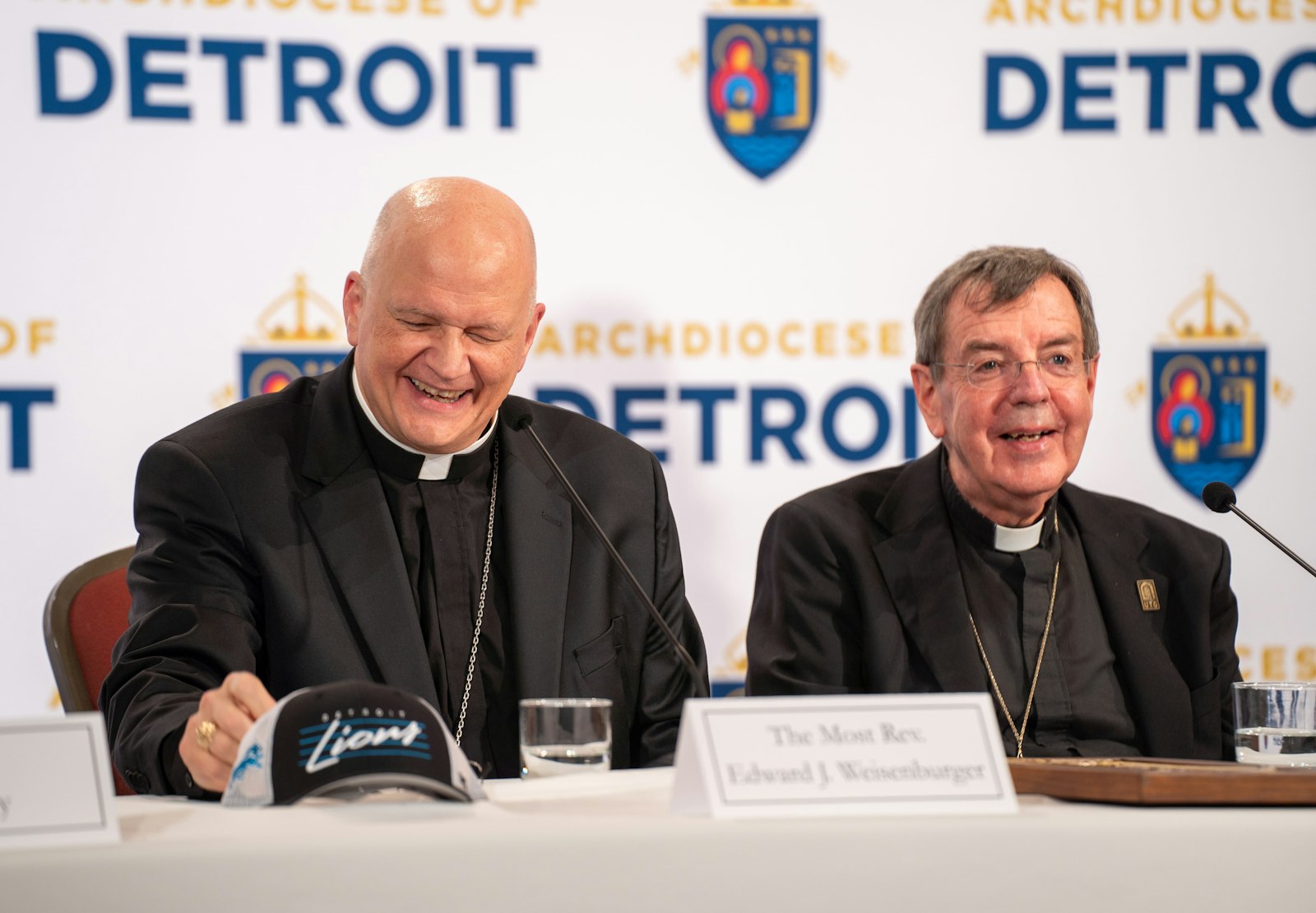 Archbishop-elect Weisenburger shares a laugh with Archbishop Vigneron, who gifted the new archbishop-elect with a Detroit Lions hat, along with a Pewabic tile with an image of Blessed Solanus Casey, at an introductory press conference Feb. 11.