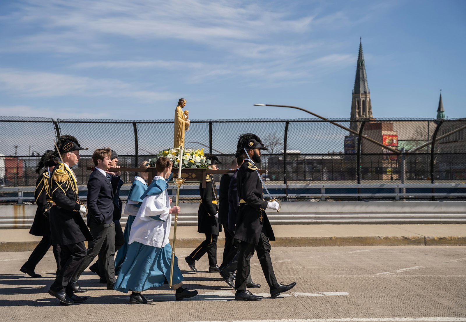 A procession with a relic and statue of St. Joseph continues over an overpass with the steeple of St. Joseph Shrine visible in the background.