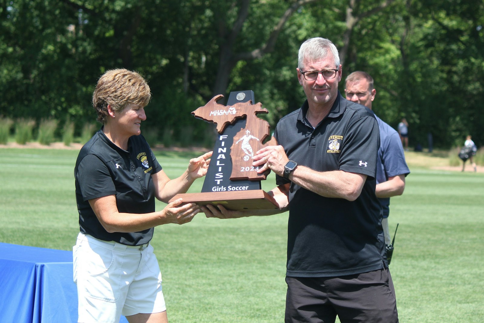 Clarkston Everest Collegiate/Bloomfield Hills Academy of the Sacred Heart soccer coach Richard Cross accepts the state runner-up trophy from Everest athletic director Ann Lowney after the Mountaineers fell to Kalamazoo Christian, 5-0, at Michigan State University.